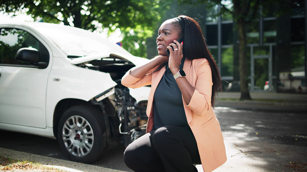 A woman talking on the phone in front of a white vehicle with damage to the front-end